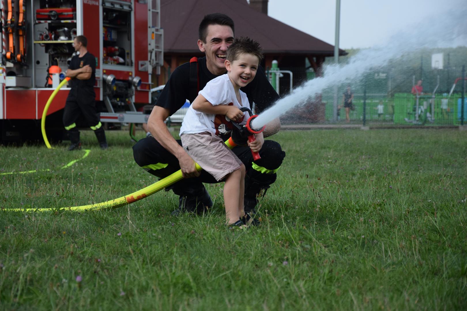 Zdjęcie przedstawiające chłopca i strażaka lejących wodę z węża